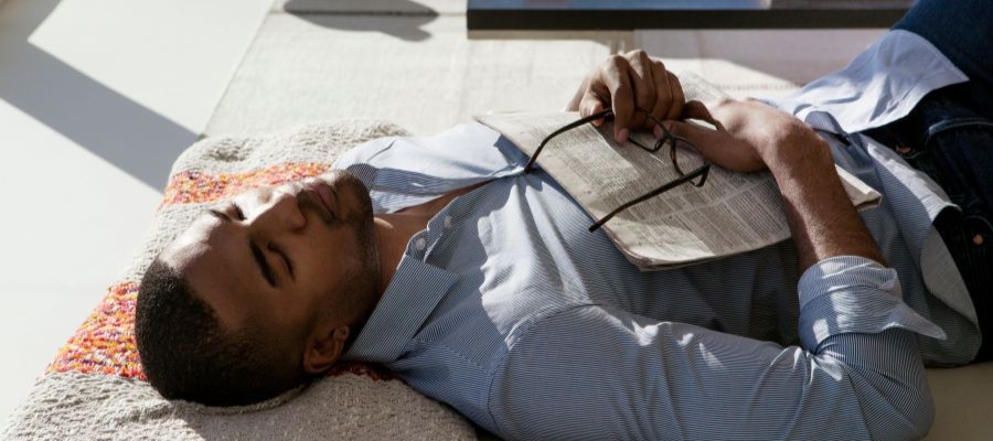 A young man is taking a nap on a rug with a newspaper and glasses on his chest. 