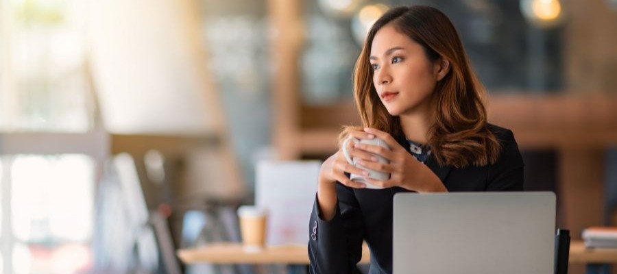 A woman sits in front of a laptop, holding a mug of coffee in her hand, staring off in the distance.