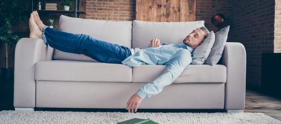 A man wearing jeans and a light blue shirt is lying on a sofa with his feet up and one arm hanging off the edge.