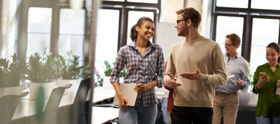 A man gestures with his hands, talking to a female coworker who holds a laptop in her hands.  There are other smiling coworkers behind them in the conference room.