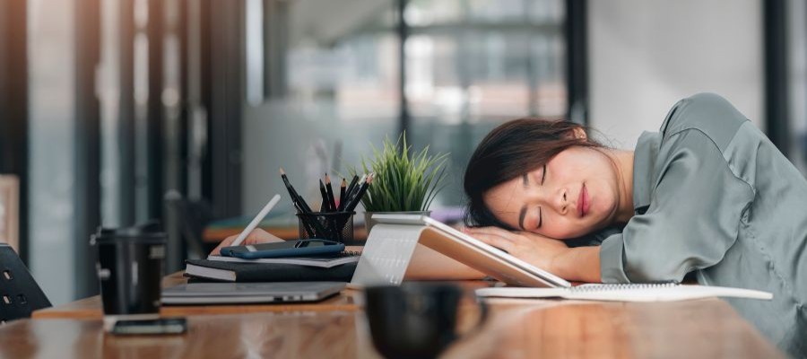 A woman is asleep with her head on her arm at her desk. It is daytime and she looks like she is taking a powernap at work.