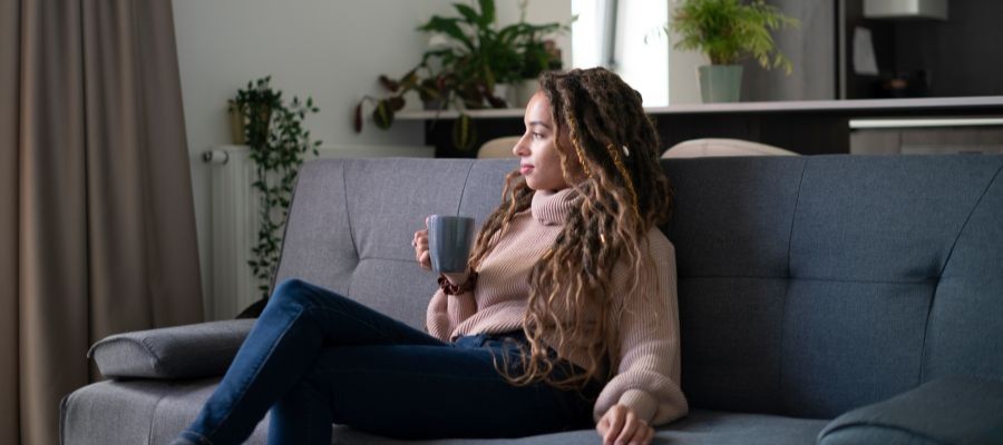 A woman sits on a grey sofa with a mug in her hand looking out the window to the side. A coffee before a nap can compound the boost in alertness you experience post-nap.