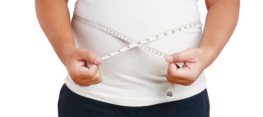 An overweight person in a white t-shirt holds a tape measure around their mid-section.