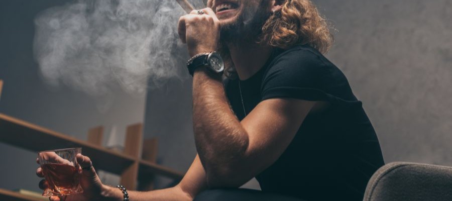 A man with long hair is holding a cigar in one hand, whiskey in the other. Both alcohol and smoking can contribute to snoring.