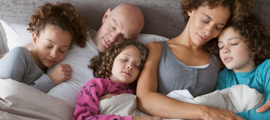 A family with two parents and 3 children of different ages is asleep in bed together. 