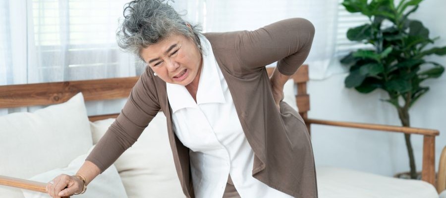 An older woman is bent over in pain. She has one hand on her lower back. 