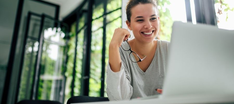 A young woman is smiling, looking at a laptop screen. She is holding glasses in her hand and sits in a room with large windows behind her, showing greenery and sunlight.