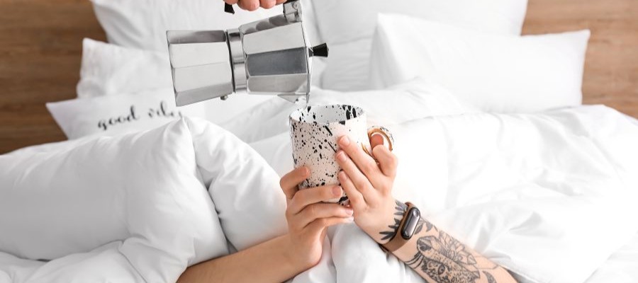 Someone pours coffee from a french press into a mug held by someone in bed under the covers. Caffeine and sleep can interrupt each other so understanding how caffeine works is key.