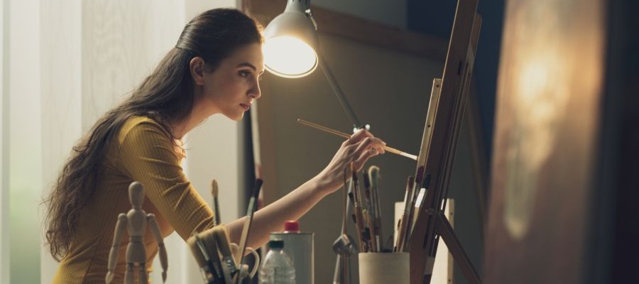 A woman is painting at an easel. She has a lamp on next to her and she is surrounded by paintbrushes and other painting supplies.