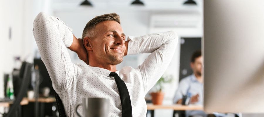 An man wearing a shirt and tie leans back at his desk with his hands behind his head, as though he is relaxed.  He has a smile on his face and he is looking off to the side.