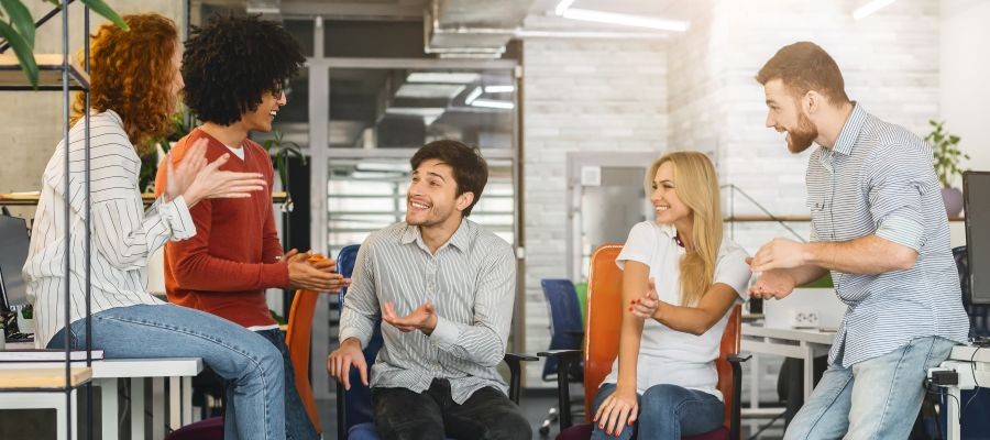 A group of 5 coworkers are gathered around, talking and laughing together. 