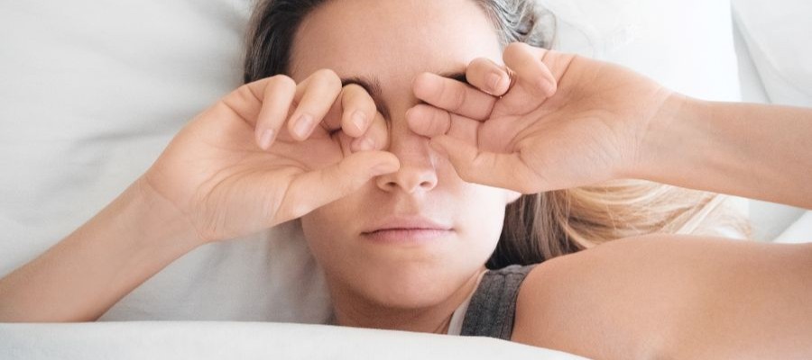A woman is lying in bed, rubbing her eyes. It looks like sunlight is coming in through a window off to the side but she does not seem ready to wake up. 