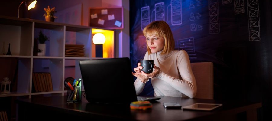 Woman sits in front a laptop in a dimly lit room. Her face is lit by the computer screen and she is holding a mag in her hands.
