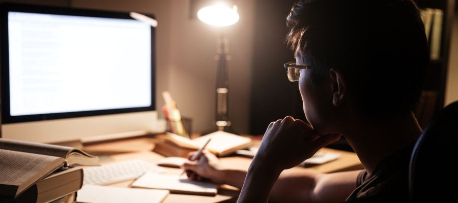 A young person sits in front of a computer, taking notes, in a dark room with just a table lamp on. 