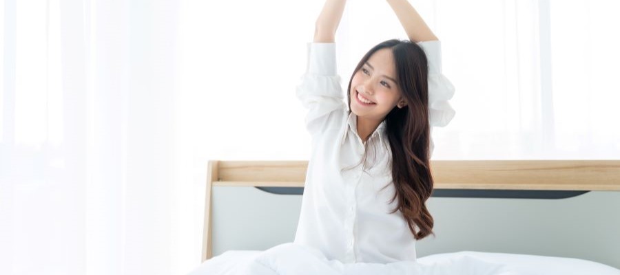 A young woman sits up in bed with her arms outstretched above her head, smiling. It looks to be daytime and she looks well-rested.