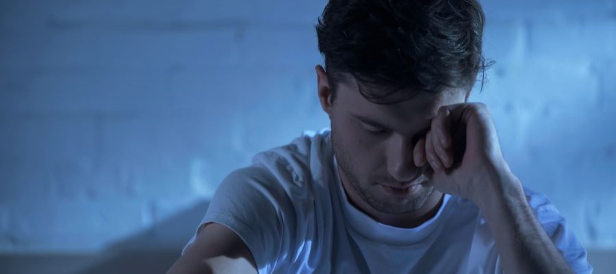 A man sits in a darkened room, rubbing his eye with one hand. There is a little light coming through as if dawn is approaching but he struggles with insomnia.