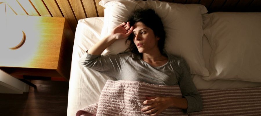 A woman is laying in bed, awake, with a bedside lamp on in an otherwise darkened room. She looks tired and stressed but not able to sleep.