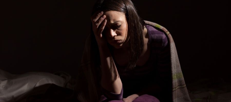 A woman is sitting in the dark with a light blanket over her shoulders. She is leaning on one hand with a frown on her face.
