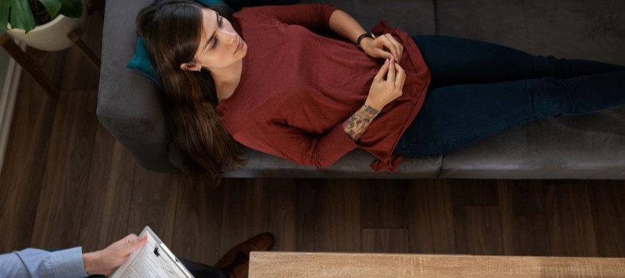 A woman is lying awake on a sofa while a therapist sits with a clipboard to the side. CBT-I can be a helpful therapy for insomnia.