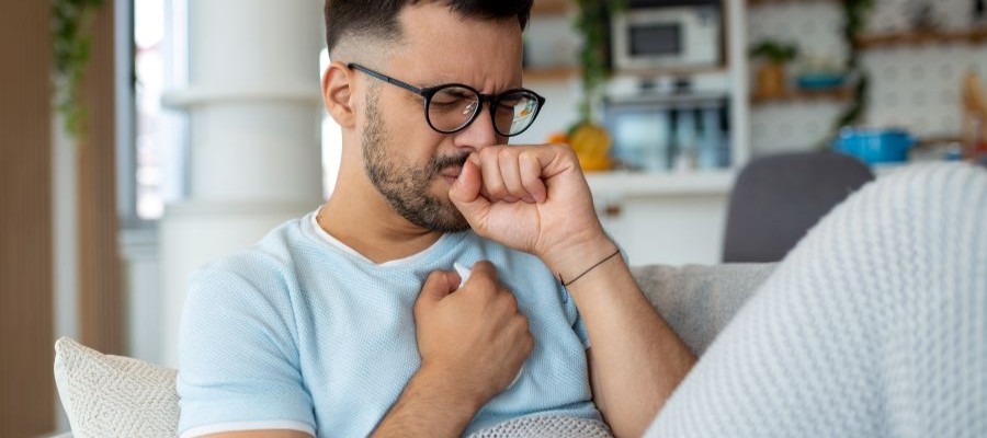 A man with glasses grimaces as he holds his chest. He is sitting on a sofa with a blanket over his bent knees. Reflux and heartburn can be a cause of maintenance insomnia.