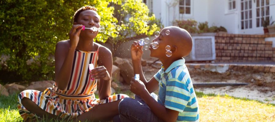 A woman and young boy sit on the grass in the backyard, blowing bubbles in the morning sunlight.