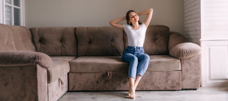 A young woman sits on a brown sofa, stretching with her arms behind her head. Her eyes are closed, there is a slight smile on her face and she looks well rested.
