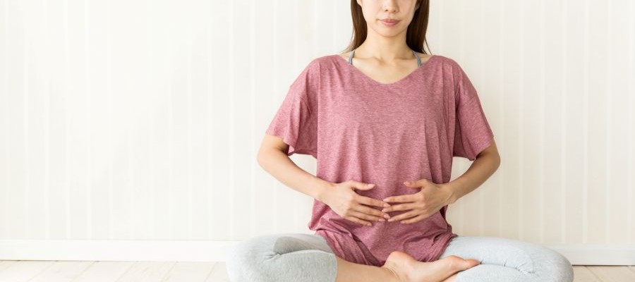 A woman sits cross-legged on the floor with her hands over her abdomen, practicing breathing exercises.