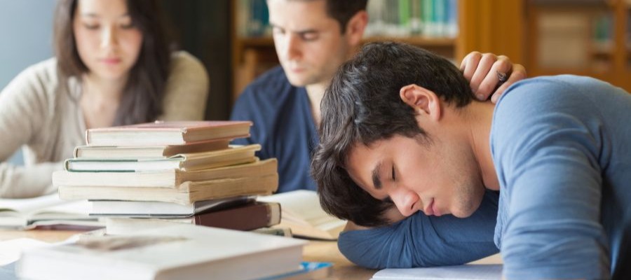 A young man is asleep at a table with his head on one arm. He is surrounded by a stock of books and notebooks and there are two people studying in the background.