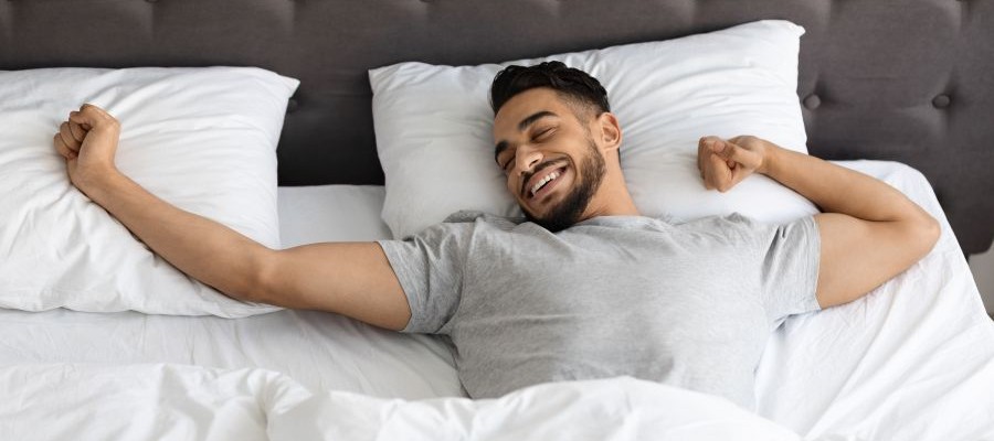 A man stretches in bed. His eye are closed but he has a smile on his face.