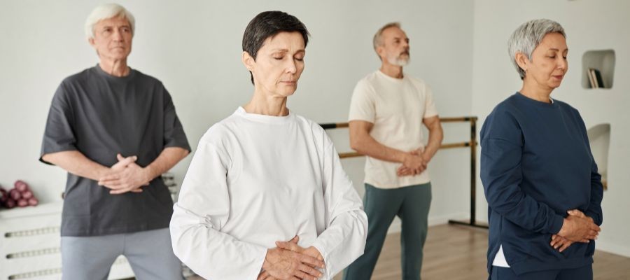 Four older adults stand with eyes closed in a workout studio, practicing breathing for relaxation.