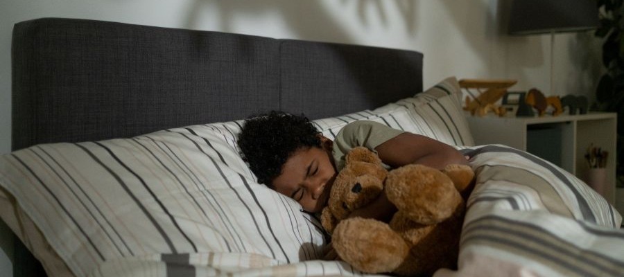 A young child is curled up in bed with a brown teddy bear. They have a frown on their sleeping face as if they are having a nightmare. There are shadows of creepy hands on the wall above their head.