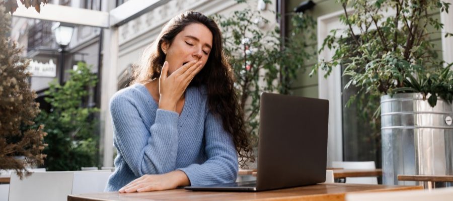 A woman with long brown hair sits outside at a sidewalk table in front of a laptop.  She is yawning with her eyes closed.