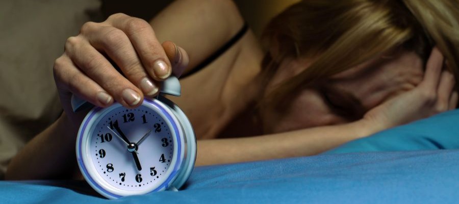 A woman is in bed, one hand snoozing a small alarm clock that reads 5:55. She is on her side with her face pressed into her other hand and the pillow.  