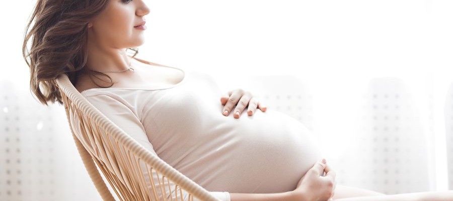A pregnant woman sits reclined on a chair holding her hands on her belly.