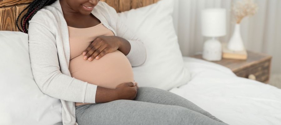 A pregnant woman sits on a bed smiling down as she holds her hands protectively around her belly.