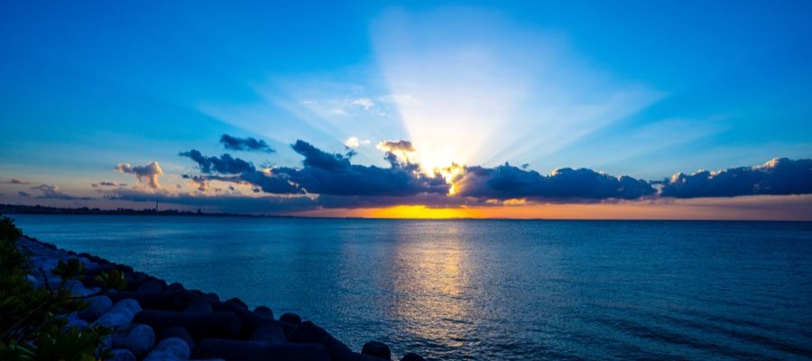 Image of a sunrise with the sun shining behind fluffy clouds over an expanse of water.  During Daylight Savings Time, daylight hours are extended giving people more time for 'daytime' activities.