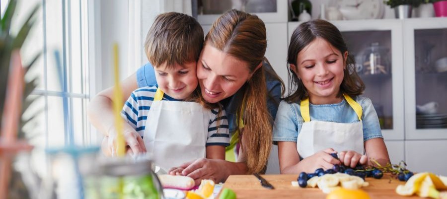 A woman is helping two young, smiling children prepare food in a kitchen.