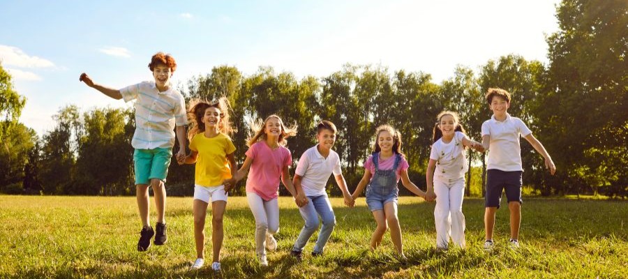 A group of 7 children are holding hand, mid big jump.  They are standing in a grassy field with trees on a sunny day.