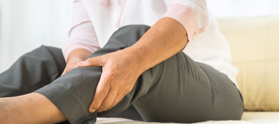 An older person sits on a bed and holds their calf as though in pain.