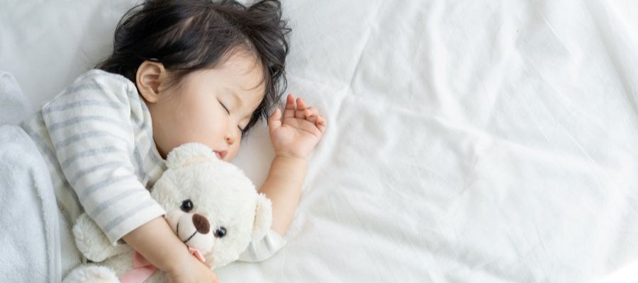A young Asian baby sleep on their side with an arm over a white teddy bear.