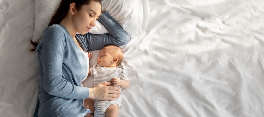 A mom sleeps on her side next to a baby on a large bed with white linens. Her hand rests on the sleeping newborn's belly and she has one arm below her head.
