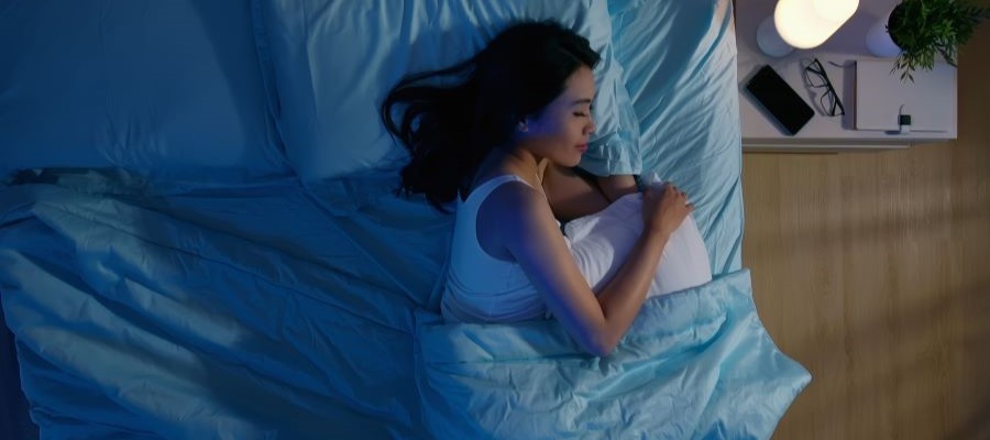Woman is asleep on her side in a bed with blue sheets. A small bedside lamp is on and it appears to be night.