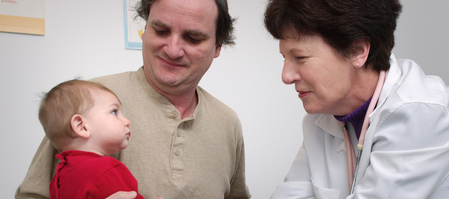 A man holds a young child on his lap in front of a female doctor who is looking at the child.