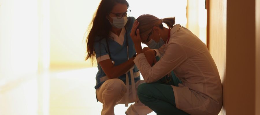Two healthcare workers crouching in a hallway, one consoling the other.
