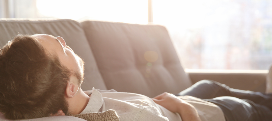 A man is asleep on a couch in the middle of the day; there is sun streaming in the window.