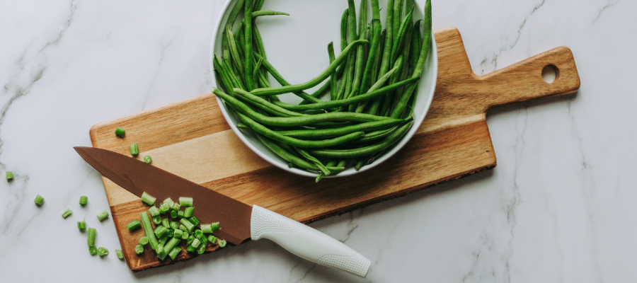 A bowl of fresh green beans sits on top of a wooden cutting board with chopped pieces of green beans.