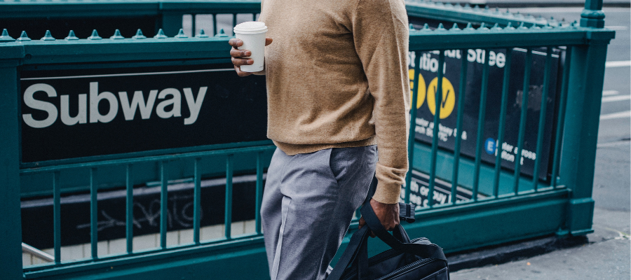 An image showing the body of a person walking past an NYC subway entrance. He is holding a coffee cup in one hand, briefcase in the other.