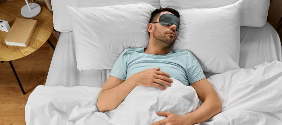 A man is asleep on his back in bed with an eye mask over his eyes.
