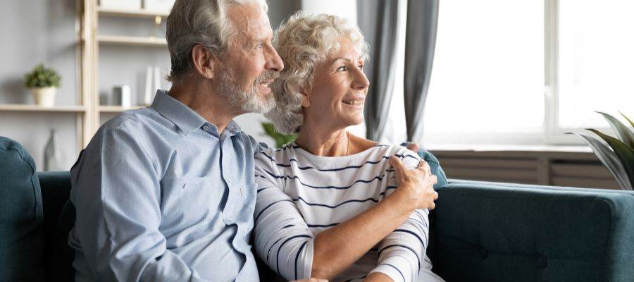 An older man and woman sit together on a couch, looking out a window. He has his arm around her and they look happy.
