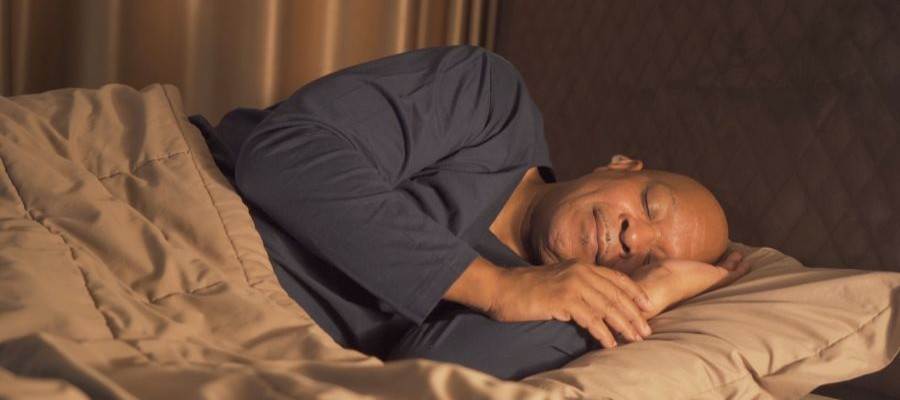A brown skinned, bald man is asleep on his side in bed with a light blanket over his body.